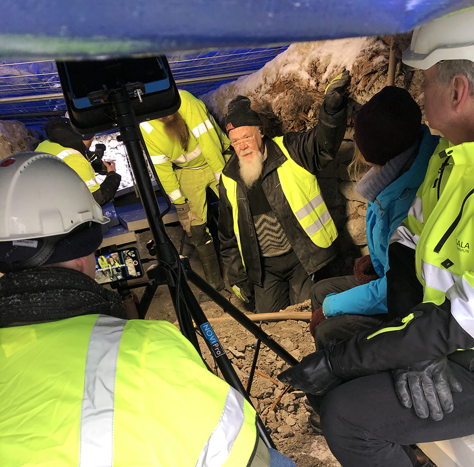 Bilden visar Leif Berggren och Jonas Wikborg som berättar för två journalister om rälsbanan som har grävts fram. På fotot syns även två personer från projektet till höger. Gruppen människor är i det utgrävda schaktet för rälsbanan, det snöar så en blå presenning har lagts som tak.